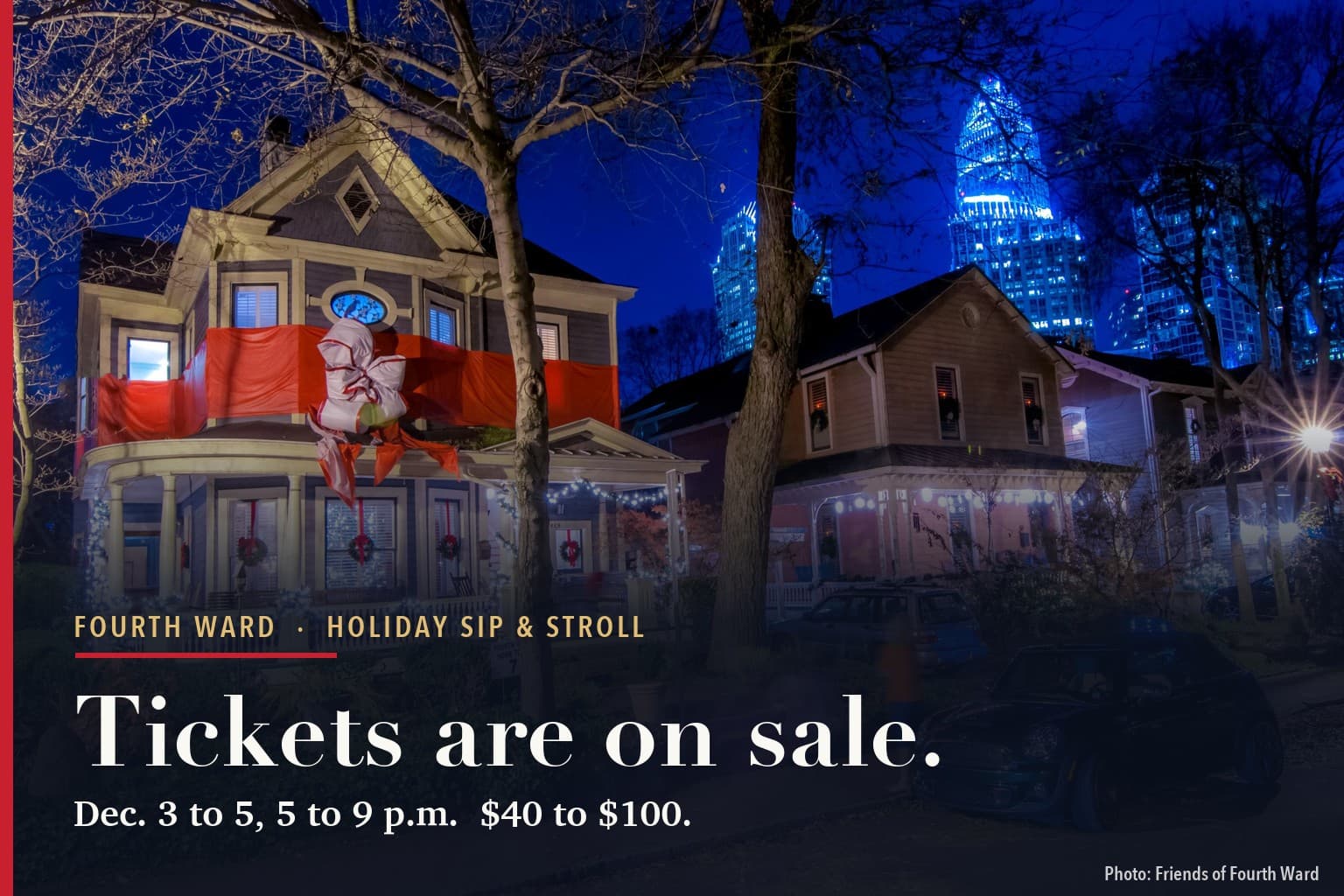Victorian houses in Historic Fourth Ward at night, decorated for the holidays with wreaths, porch lights and a red bow wrapped around a balcony, with Uptown Charlotte towers lit blue behind them.
